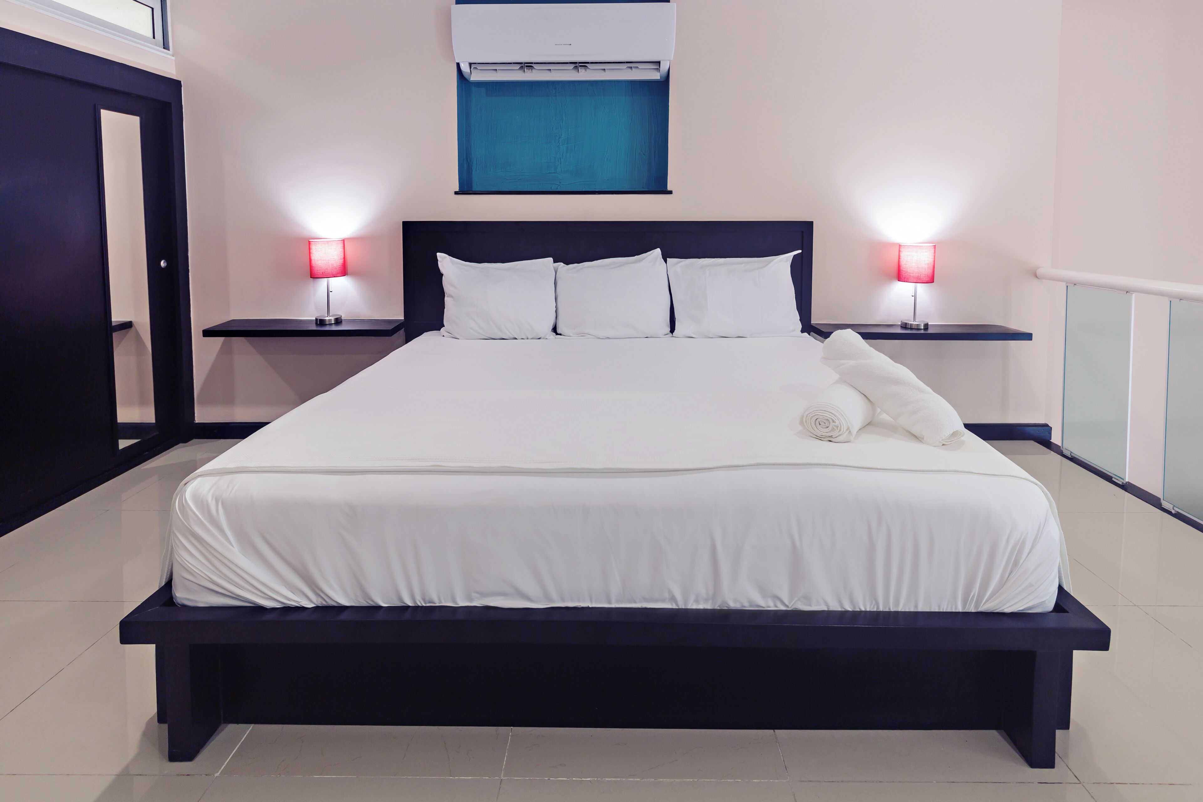 A neatly made bed with white linens, two bedside lamps, a wall-mounted air conditioner, and a window with blue curtains.