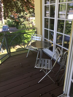 En träveranda med metallstolar och ett bord, med utsikt över en trädgård och en gata.