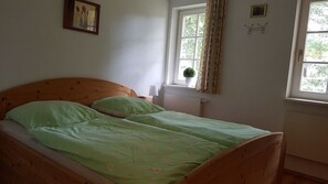 En säng för en person med en träram som huvudgavel, ett fönster med gardiner, en liten krukväxt och en tavla som hänger på väggen.