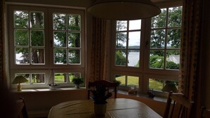 A room with large windows offering a view of a lake and greenery. There is a dining table with chairs and a small ship model on the windowsill.