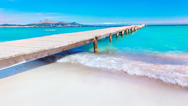A wooden pier extending into a turquoise sea with clear skies and a distant mountain. - NEW Apt RELAX HOME 150 meters from the beach, WIFI and Pool (Port d'Alcúdia)