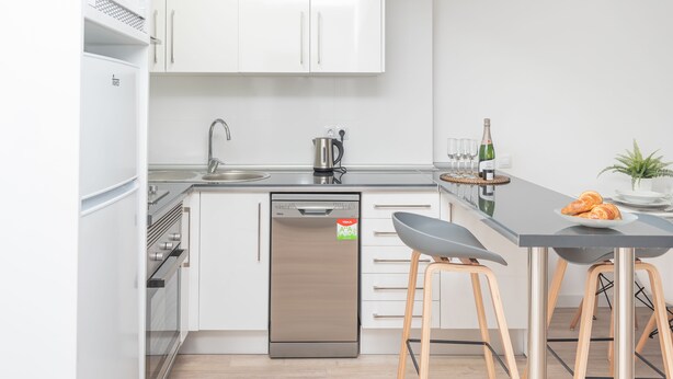 A modern kitchen with white cabinets, a dishwasher, a sink, and a bar area with stools and a bottle of champagne. - NEW Apt RELAX HOME 150 meters from the beach, WIFI and Pool (Port d'Alcúdia)