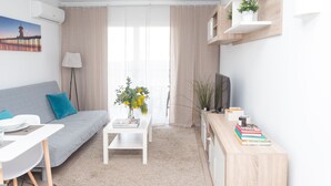 A modern living room with a grey sofa, white coffee table, and a TV on a wooden stand. There are plants on shelves and a painting on the wall. - NEW Apt RELAX HOME 150 meters from the beach, WIFI and Pool (Port d'Alcúdia)
