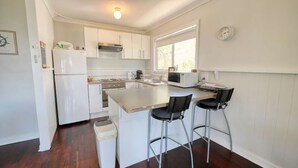 A modern kitchen with white cabinetry, a stainless steel refrigerator, a microwave, and a sink. There are two bar stools and a trash can. - Abbey Beachviews with WiFi (Abbey)