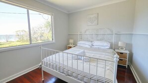 A bedroom with a white metal bed frame, two bedside tables with lamps, a window with a view of trees and a body of water, and a wall-mounted map. - Abbey Beachviews with WiFi (Abbey)