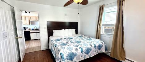 A bedroom with a bed, a ceiling fan, a window with curtains, and a view of a kitchen.