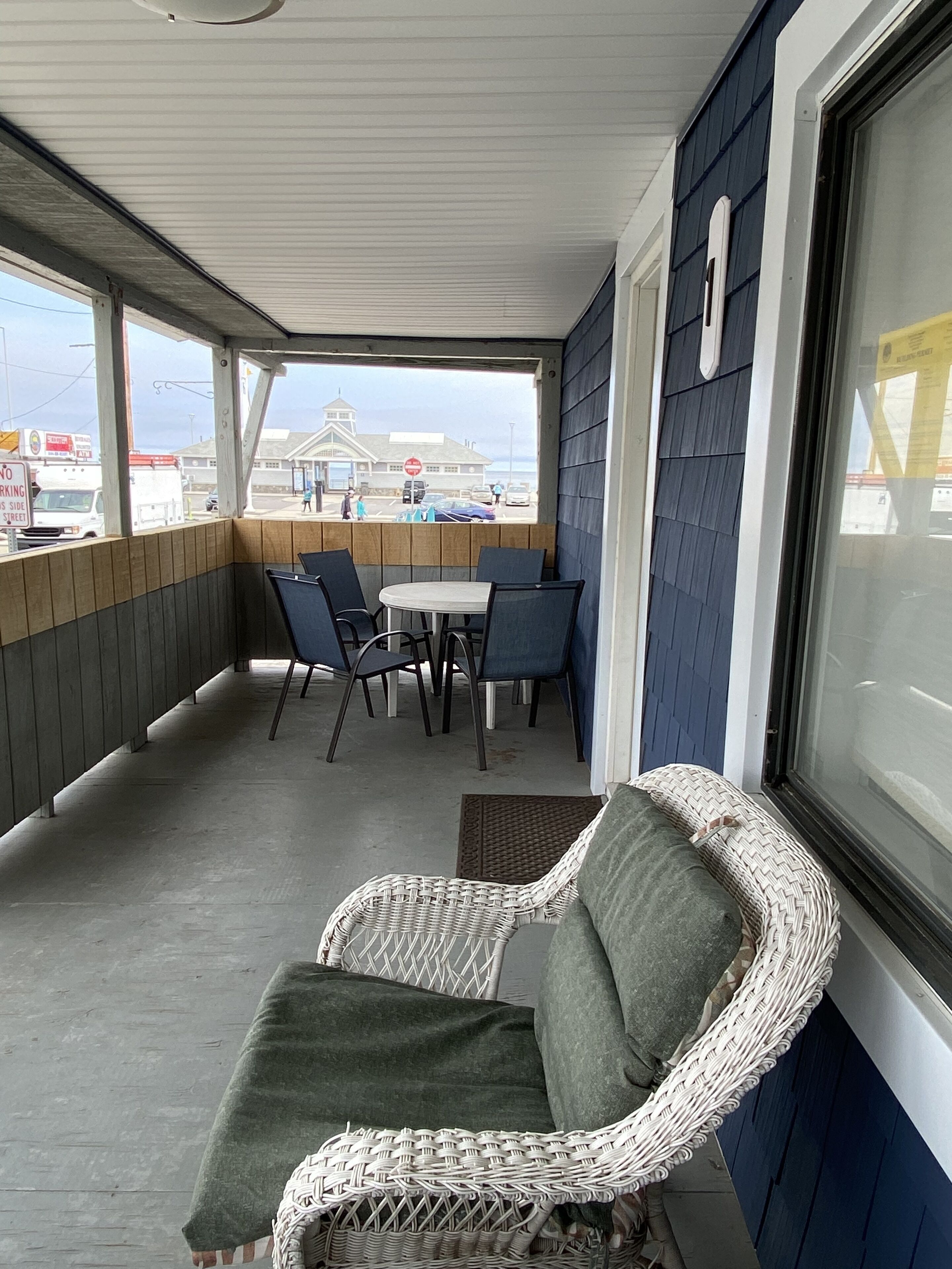 A covered outdoor seating area with wicker chairs and a table, overlooking a beach and a building with a tower.