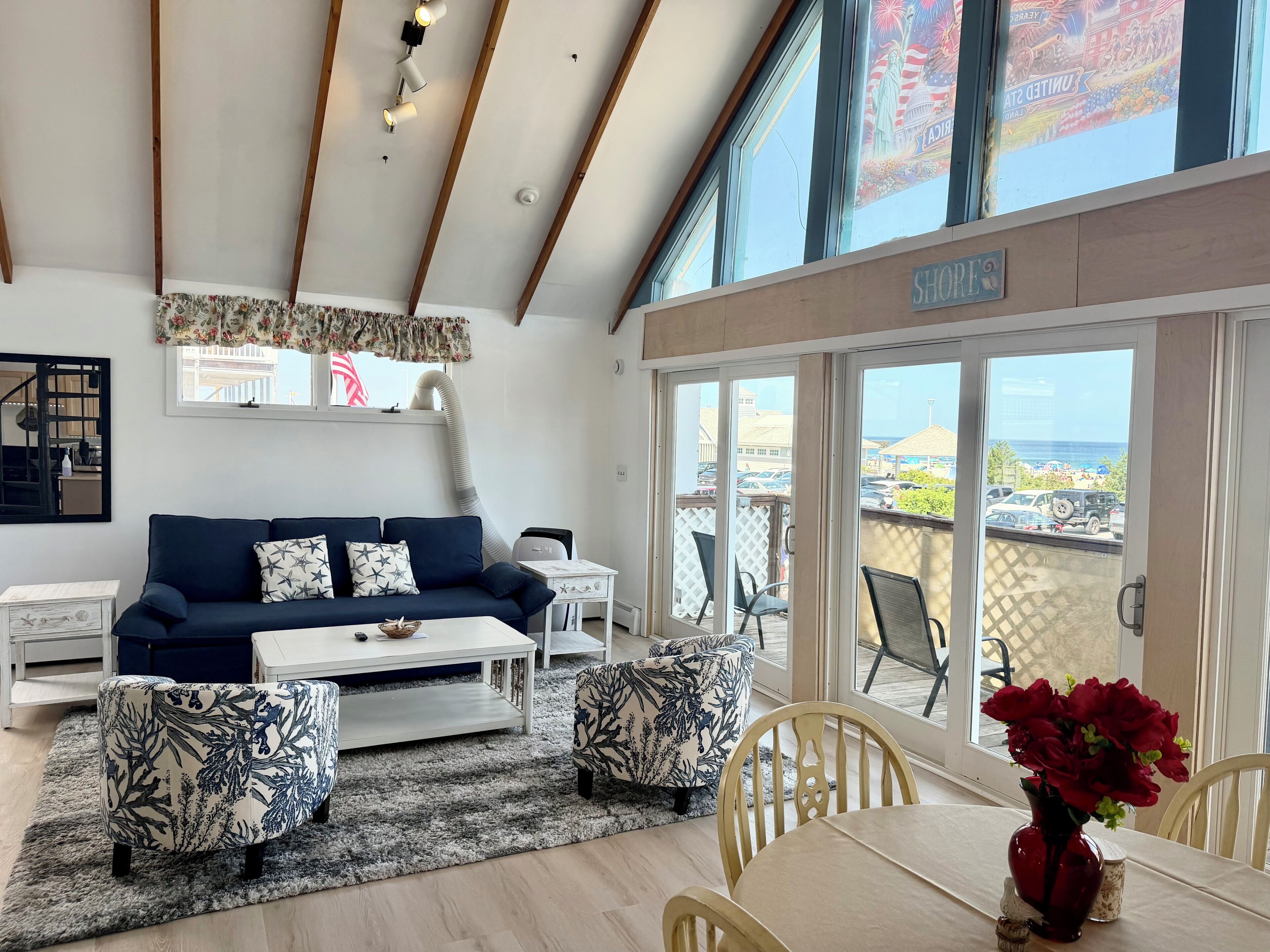 A spacious living area with a sofa, coffee table, and chairs, a dining table with a vase of flowers, and large windows offering a view of the beach and ocean.
