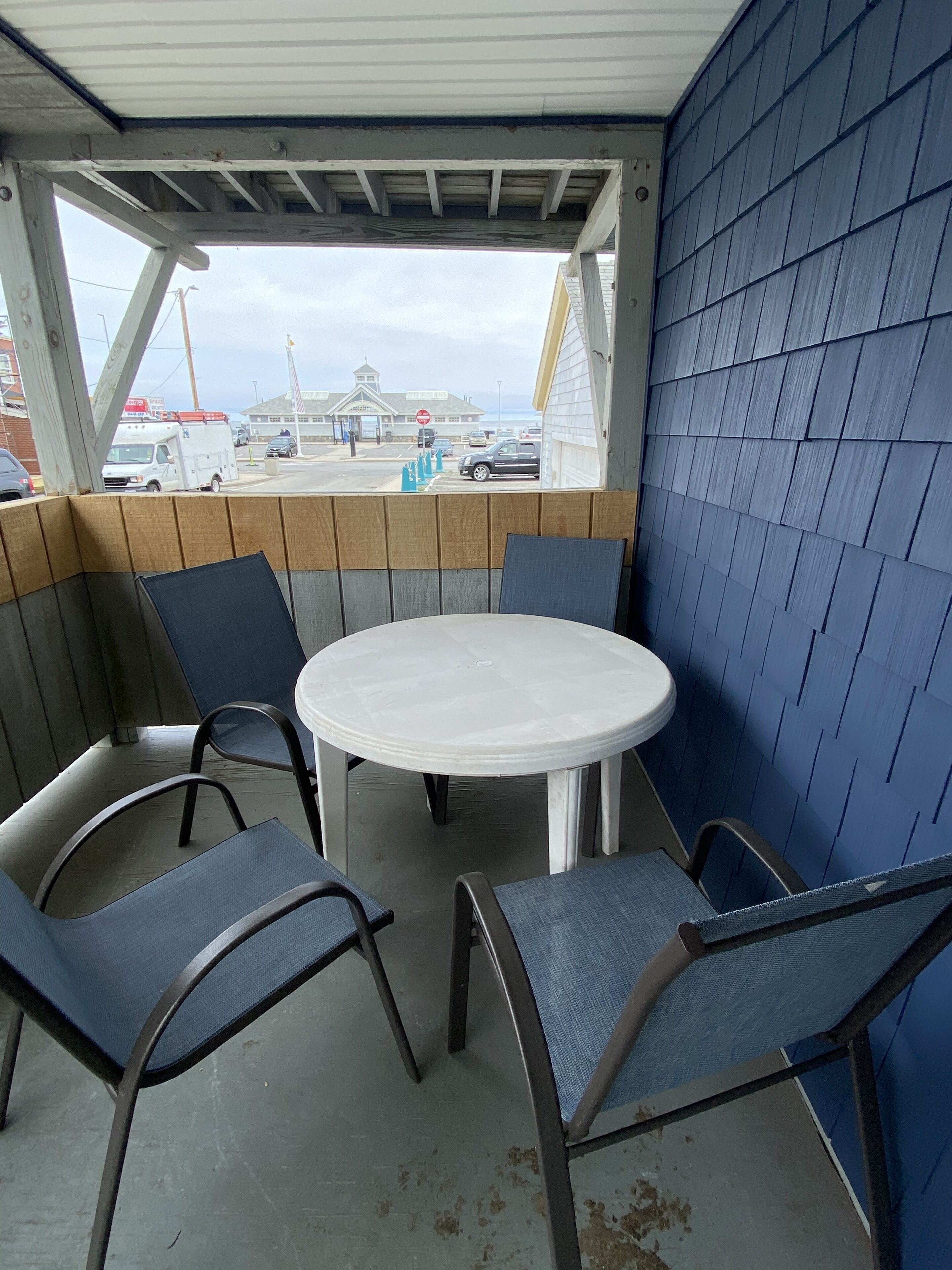 A covered outdoor seating area with a round table and four chairs, a view of a building and parked cars in the background.