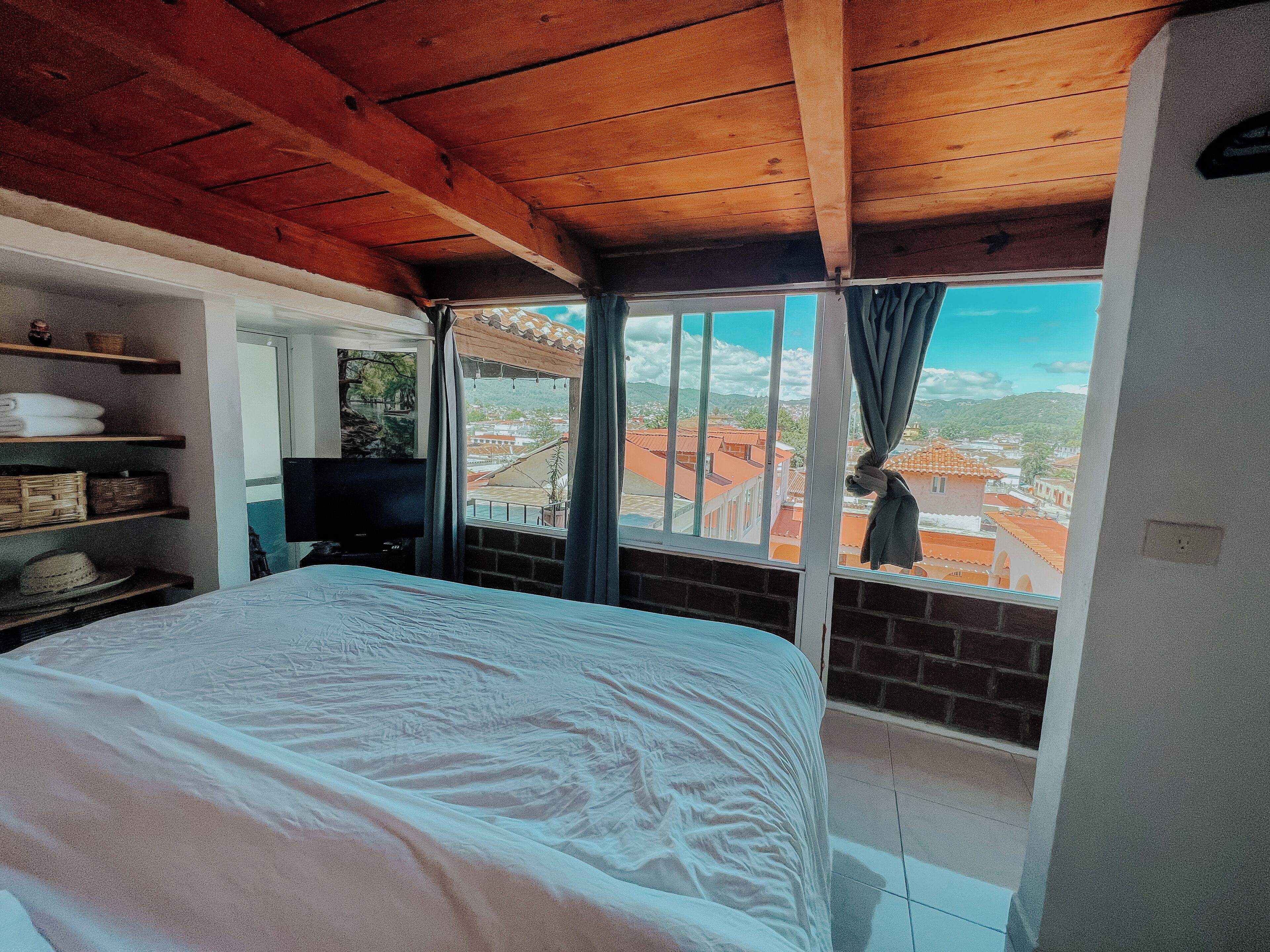 A bedroom with a large bed, wooden ceiling beams, a TV, and a window with a view of buildings.