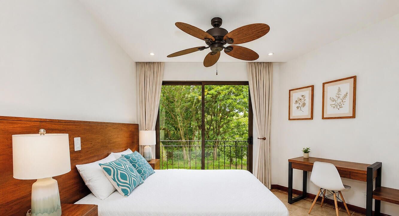 A hotel room with a bed, a desk, a chair, a ceiling fan, and a balcony with a view of greenery.