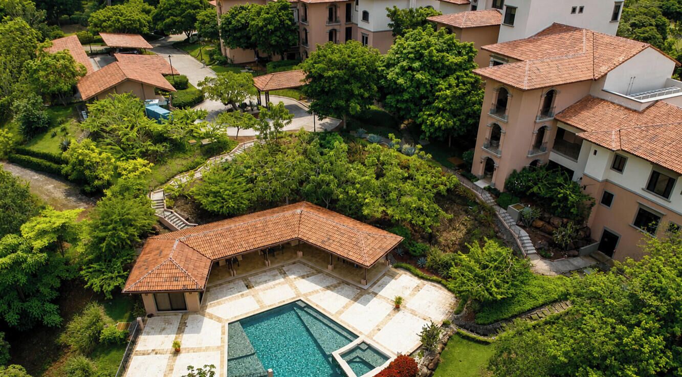 An aerial view of a residential complex with multiple buildings, a swimming pool, and surrounding greenery.