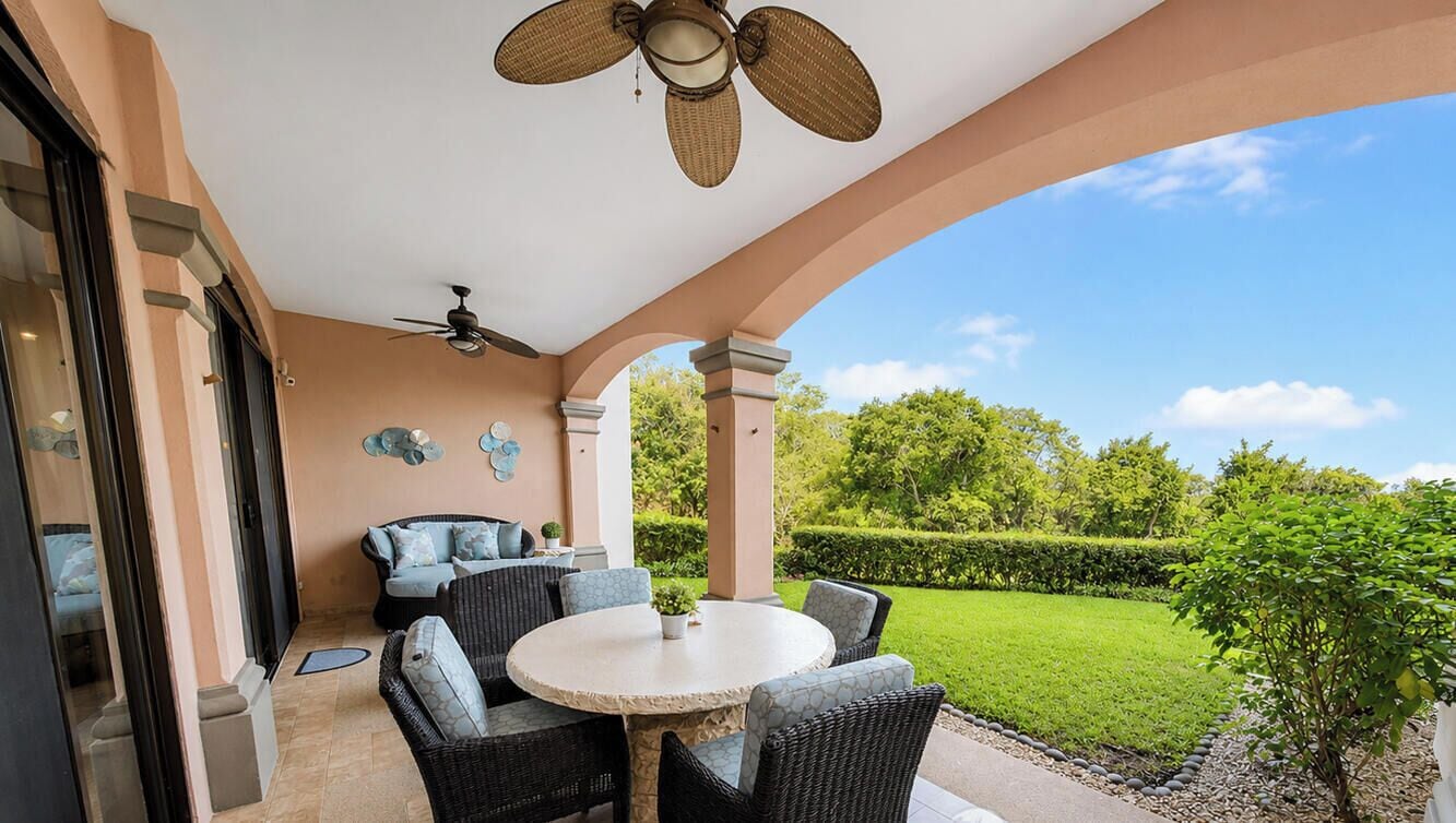A covered patio with a round table and chairs, a ceiling fan, and a view of a well-maintained garden.