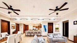 A spacious living area with a dining table, two sofas, ceiling fans, and a kitchen in the background.