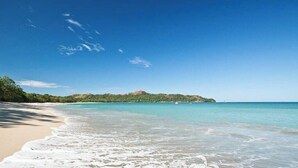 A beach with clear water, a sandy shore, and a distant hill.