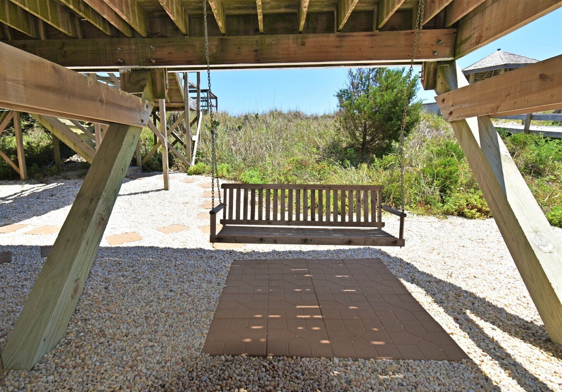 A wooden bench under a pergola with a gravel path and greenery in the background.