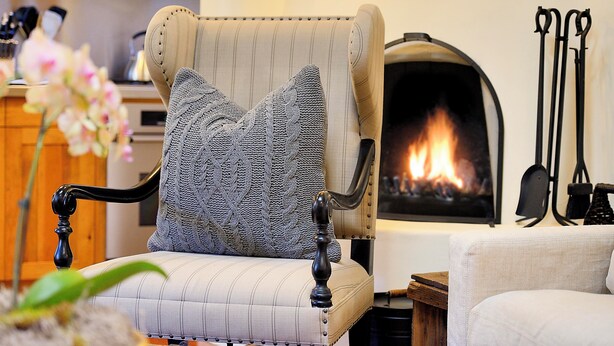 A chair with a knitted pillow, a fireplace, and a wooden table in a cozy room.