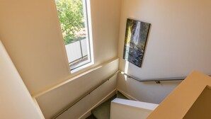 Una escalera estrecha con barandilla, una ventana con vistas a la vegetación, y un cuadro en la pared.