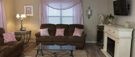 A living room with a brown sofa, a glass coffee table, a fireplace, and a ceiling fan.