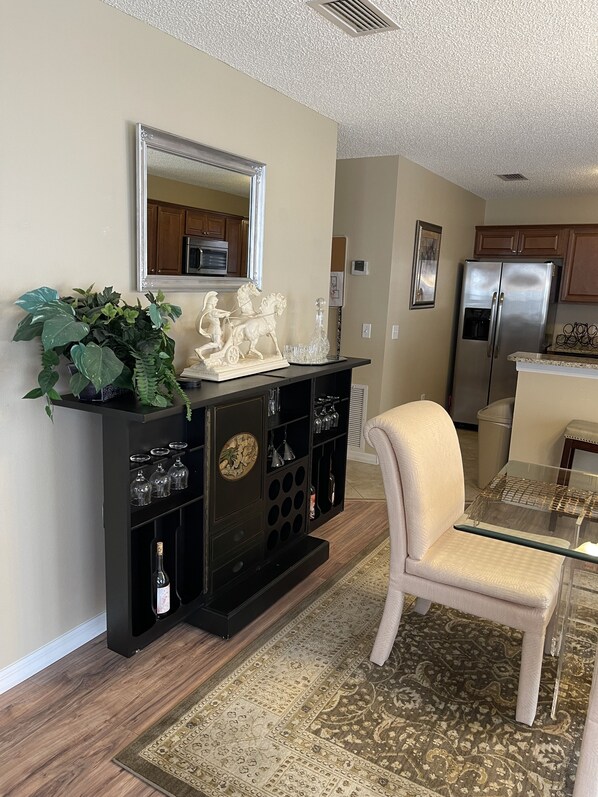 A living room with a dark wooden entertainment center, a beige armchair, a glass coffee table, a patterned rug, and a kitchen in the background. - Marilyn’s Romantic Getaway (The Villages)