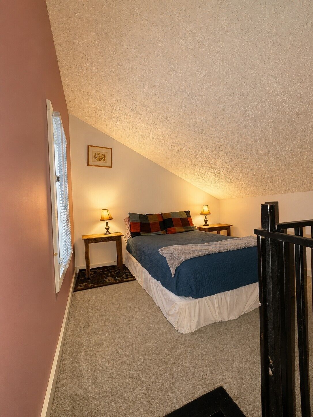 A bedroom with a bed, two bedside tables, a window with blinds, a picture on the wall, and a staircase.