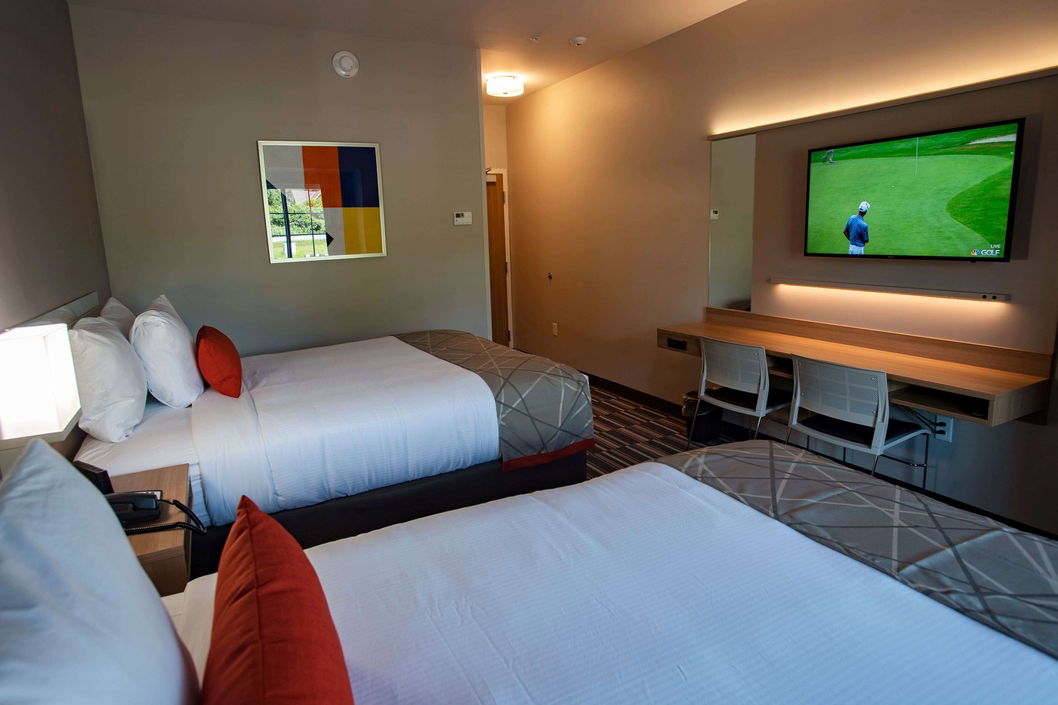 A hotel room with two beds, a TV displaying a golf game, a desk with chairs, and a wall-mounted TV.