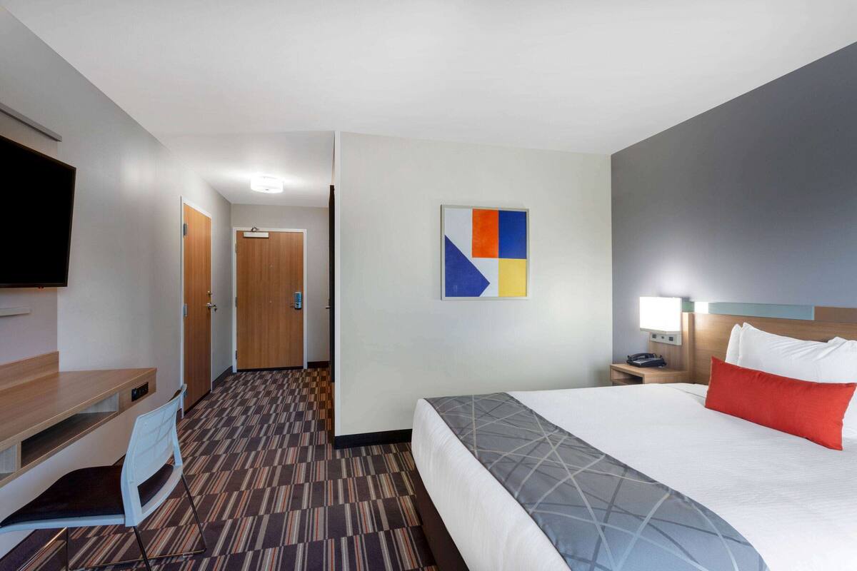 A hotel room with a bed, a desk, a chair, a TV, a nightstand, and a wall art piece.