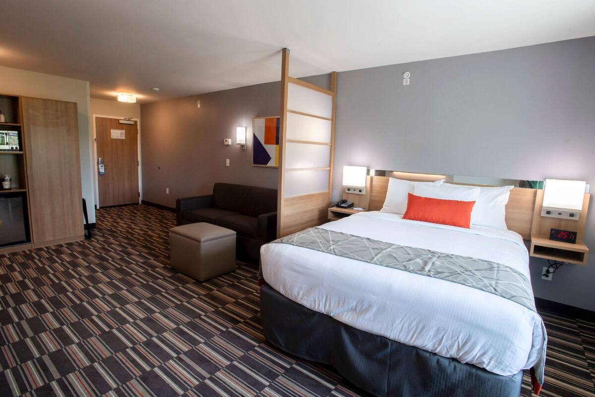 A hotel room with a large bed, a desk, a chair, a wardrobe, and a small kitchenette.