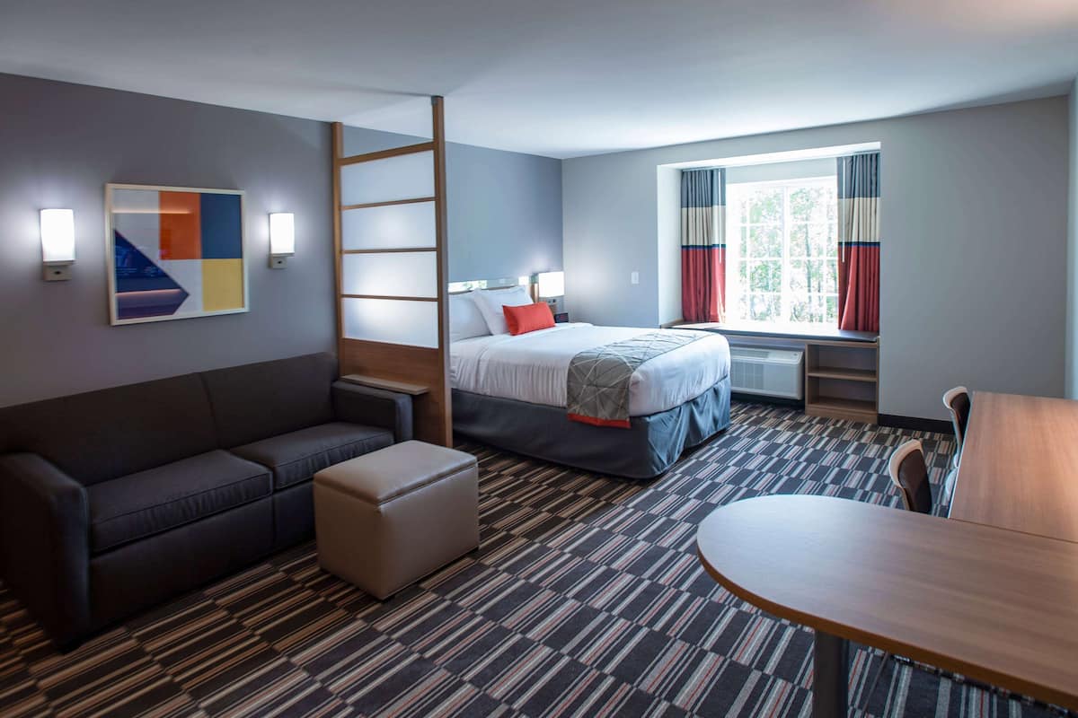 A hotel room with a bed, a sofa, an ottoman, a table, and a window with curtains.