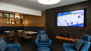 A modern lounge area with a flat-screen TV displaying a hockey game, comfortable seating, a fireplace, and a wooden panel wall. - Microtel Inn & Suites by Wyndham Amsterdam (Amsterdam)