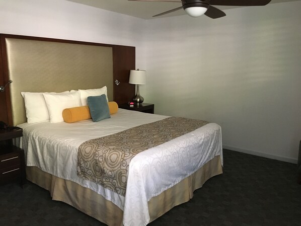 Una habitación de hotel con una cama grande, un ventilador de techo, una mesita de noche con lámpara y una puerta.