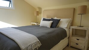 Una cama bien hecha con un edredón gris oscuro y almohadas blancas, una manta de color claro, y una mesita de noche con una lámpara y un cajón pequeño.
