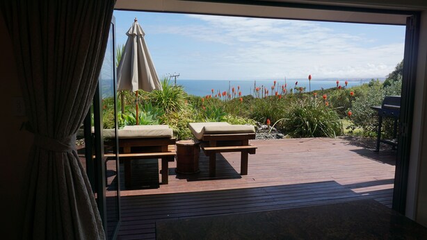 Una terraza de madera con sillones y una mesa, con vistas a un paisaje costero con vegetación y un cielo despejado.