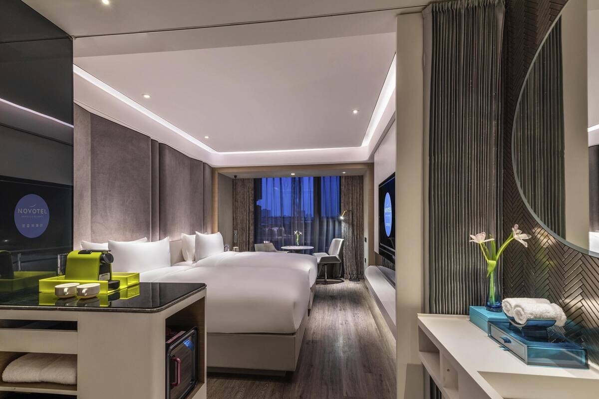 Une chambre d’hôtel moderne avec un grand lit, une télévision, un bureau avec une lampe et une vue sur la ville au crépuscule.