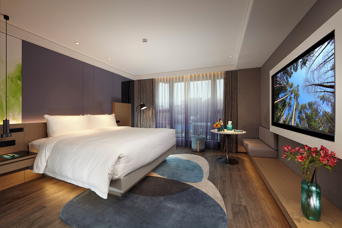 Une chambre d’hôtel moderne avec un grand lit, un téléviseur  à écran plat, une petite table et un vase rempli de fleurs.