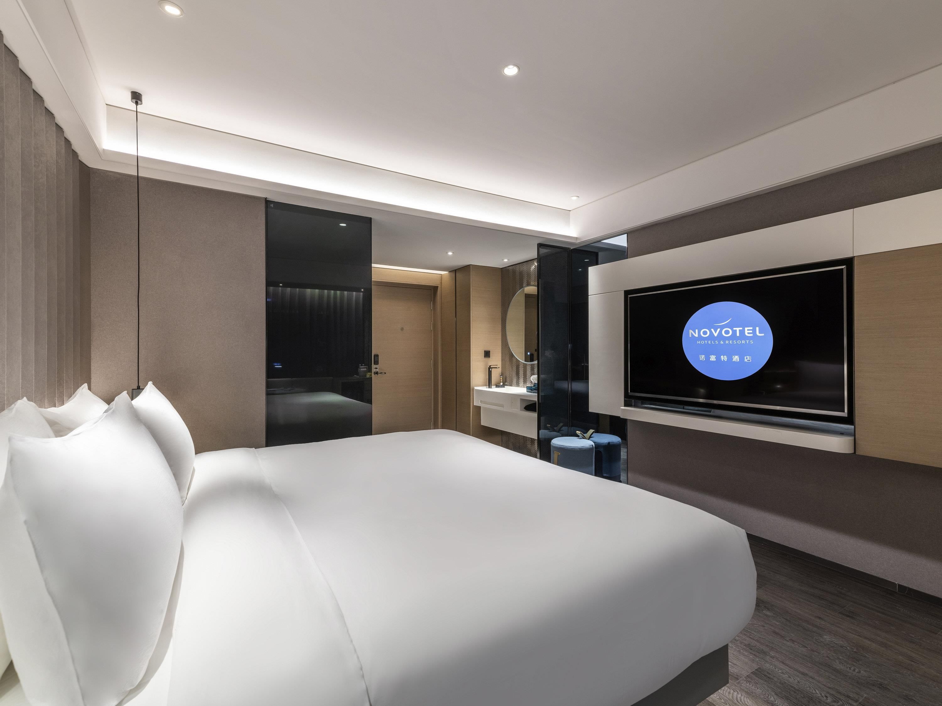 A modern hotel room with a large bed, a TV mounted on the wall, and a bathroom with a mirror and sink.