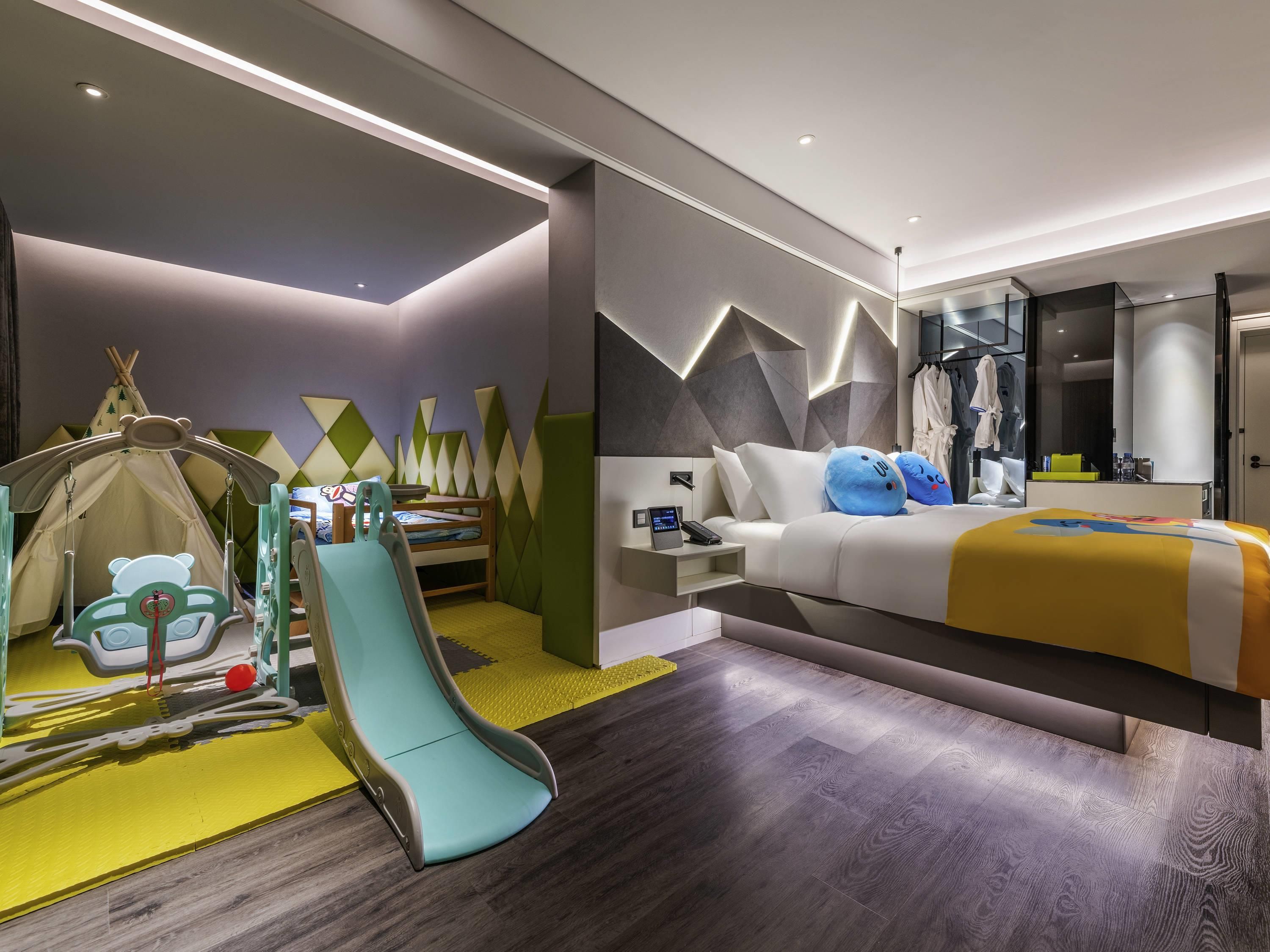 A modern hotel room with a bed, a desk, a chair, a play area, and a wardrobe.