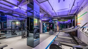 Une salle de sport moderne équipée de matériel d’exercice, de miroirs et d’un écran numérique affichant 19 h 01.