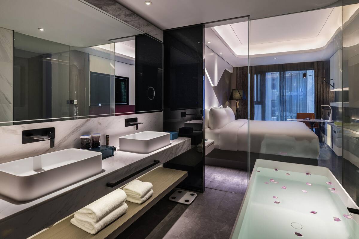 Une chambre d’hôtel moderne dotée d’un grand lit, de deux lavabos, d’une baignoire et d’un grand miroir.