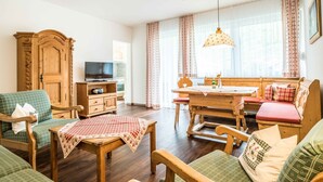 Ein gemütliches Wohnzimmer mit einem Holzschrank, einem Fernseher, einem Esstisch mit Bänken und einem Fenster mit Vorhängen.