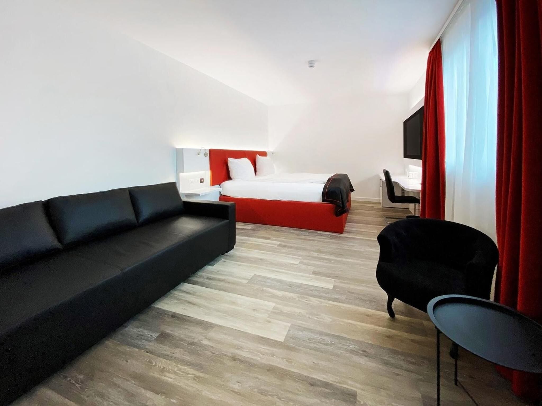 A hotel room with a black sofa, a bed with red headboard, a small black chair, and a TV on the wall.