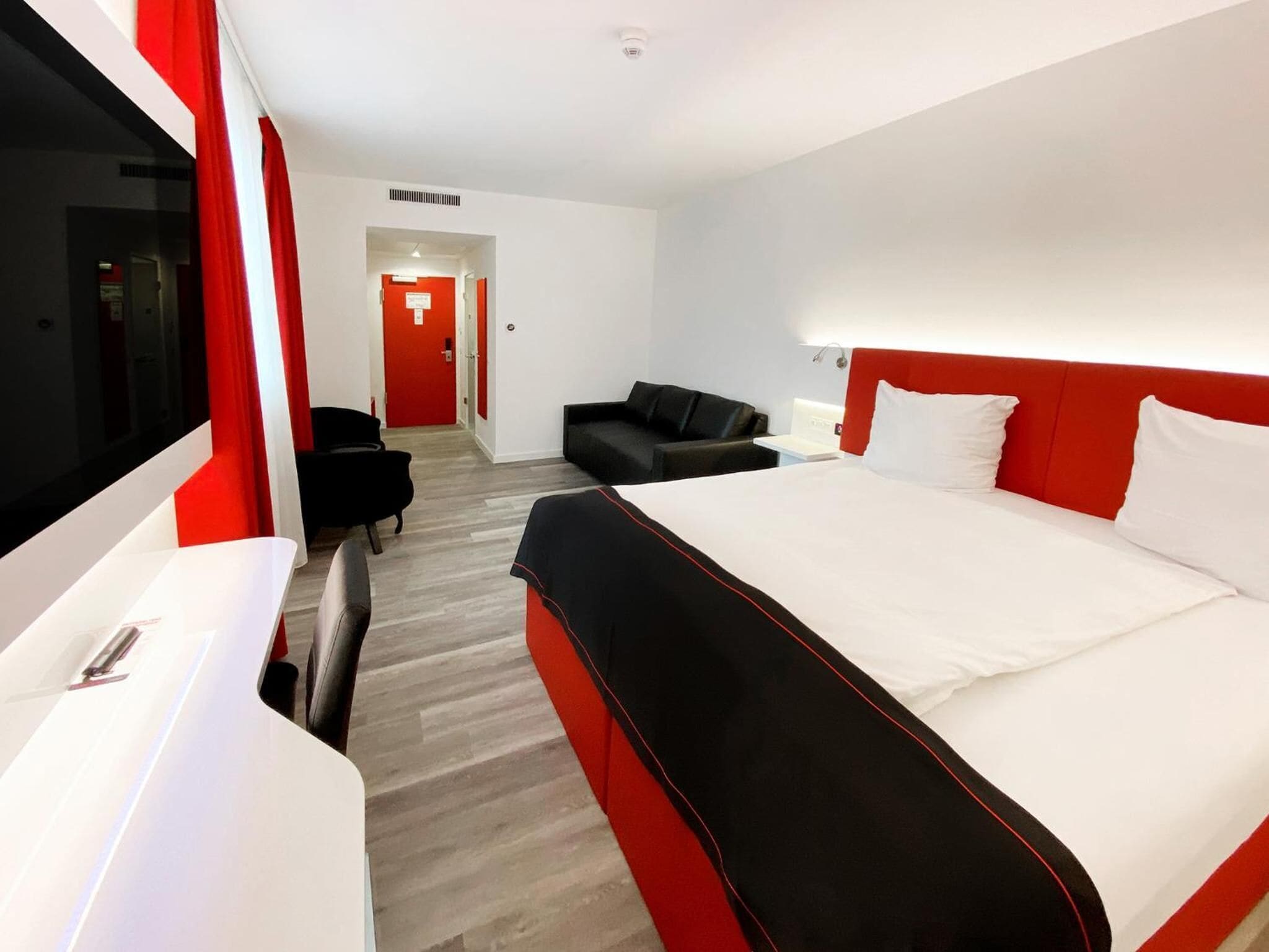 A modern hotel room with a large bed, a desk, a chair, and a sofa.