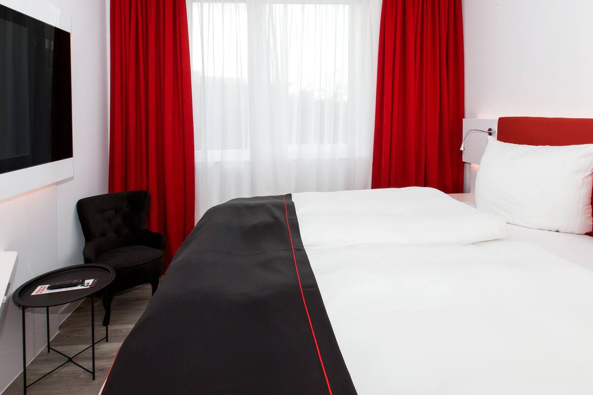 Ein Hotelzimmer mit einem großen Bett, roten Vorhängen, einem schwarzen Sessel, einem kleinen Tisch und einem an der Wand montierten Fernseher.
