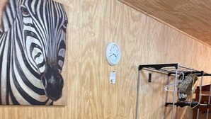 A room with a wooden wall, a zebra painting, a clock, a metal shelf unit, and a dark wooden table with benches. - Tiny House close to Buffalo Point (Saint Joe)
