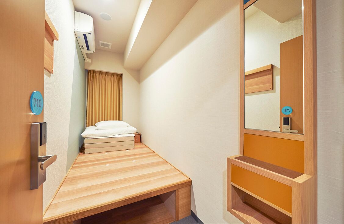 Een kleine kamer met een bed, een spiegel, een klein plankje en een deur met een nummerbord.