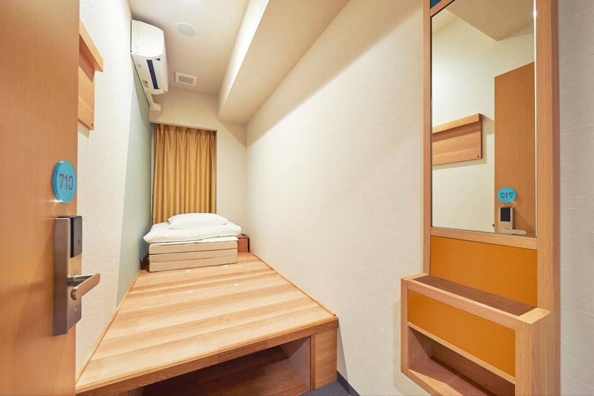 Ein kleines Zimmer mit Bett, Spiegel, einer kleinen Ablage und einer Tür mit einer Nummer.
