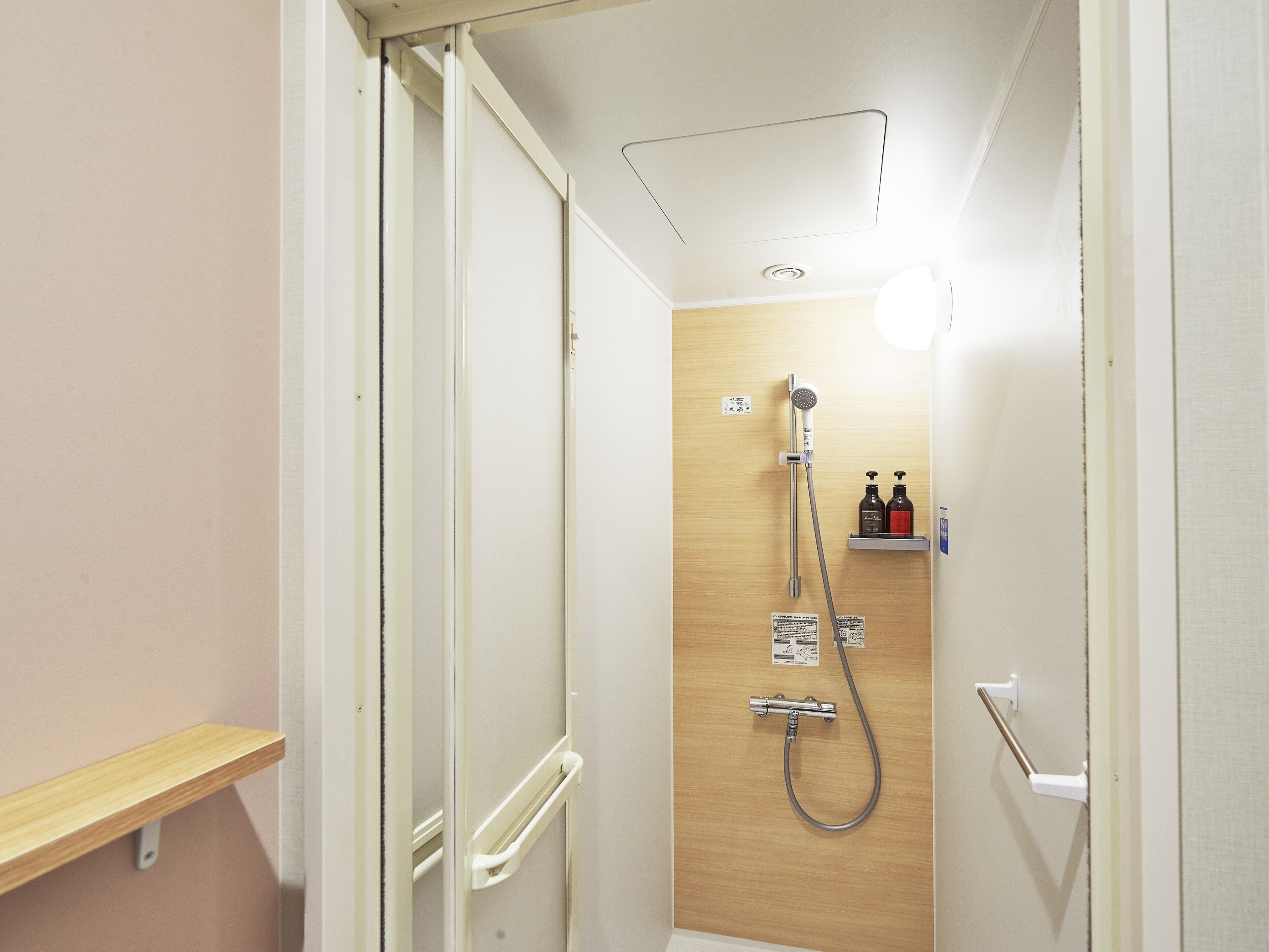 A narrow shower stall with a wooden door, a handheld showerhead, a wall-mounted faucet, and two bottles of shampoo and conditioner on a shelf.