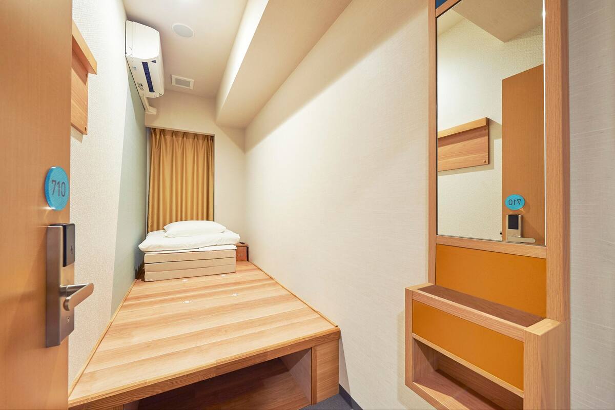 Ein schmaler Raum mit einem Bett, einer hölzernen Bank, einem Spiegel und einer Tür mit einer Nummer.