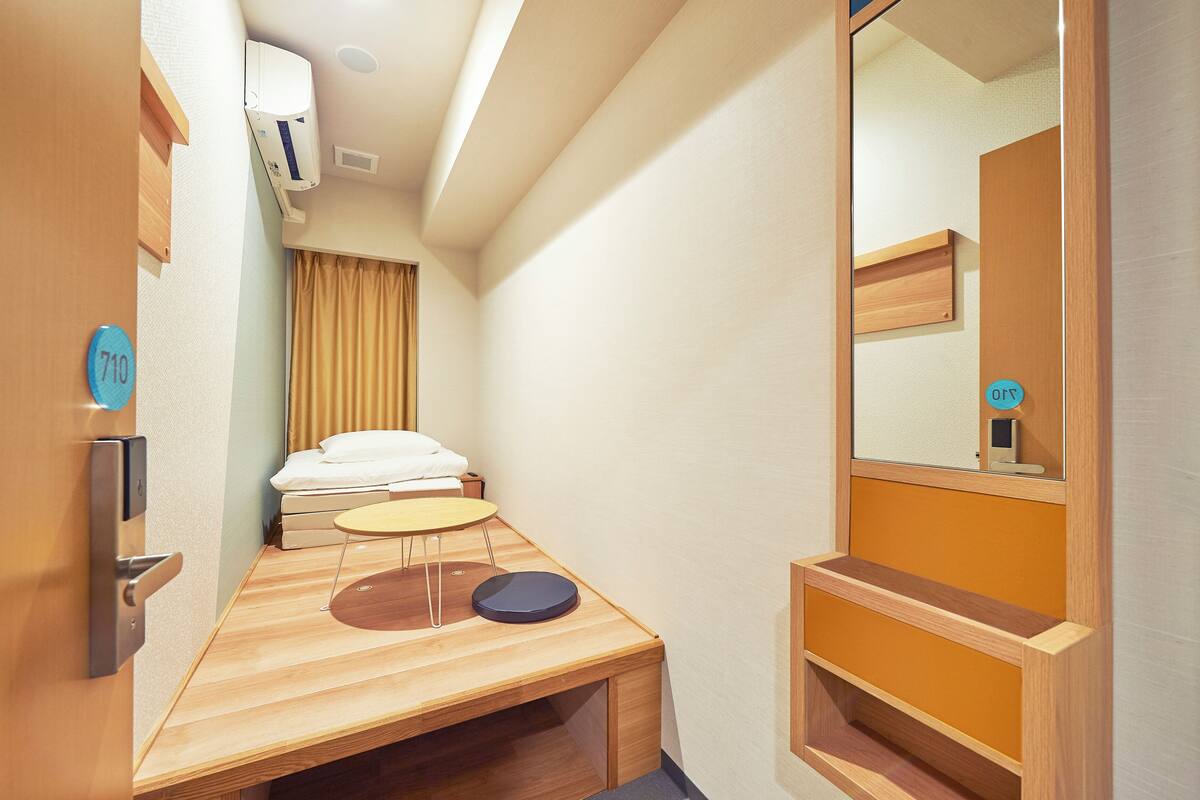Ein kleines Hotelzimmer mit Bett, Tisch, Spiegel und einer Holzbank.