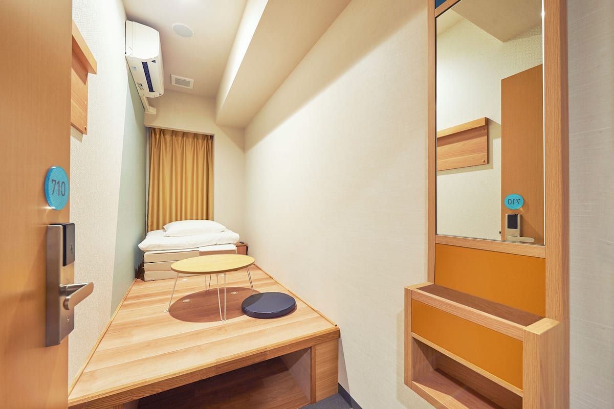 Quarto de hotel compacto com cama, uma mesinha, espelho e um banco de madeira.